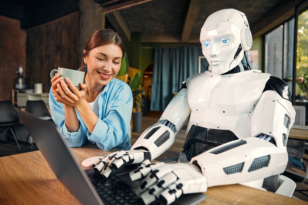 Genç bir kadın ve insansı robot, modern bir ofiste dizüstü bilgisayar başında birlikte çalışıyor; ikili, kahve eşliğinde yapay zeka destekli iş birliği yapıyor.